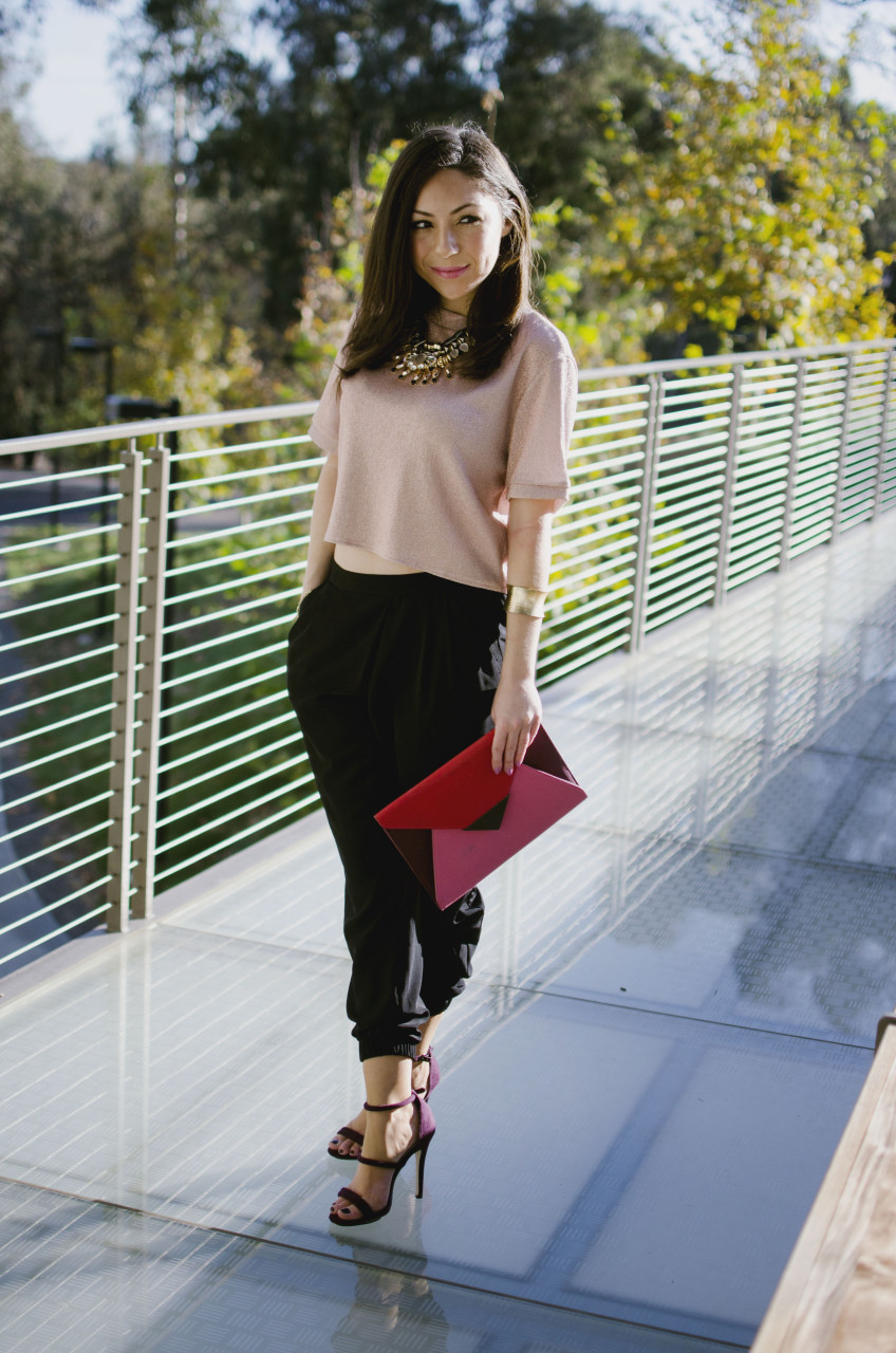 Blogger Nihan standing and showing her Holiday Party Outfit; a pink metallic top, a big statement necklace, black jogger pants, maroon highly and a color block clutch.