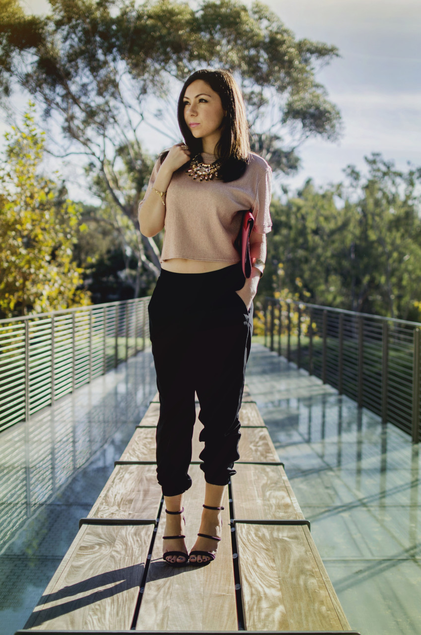 elegant holiday party look - Blogger Nihan standing and showing her Holiday Party Outfit; a pink metallic top, a big statement necklace, black jogger pants, maroon highly and a color block clutch.