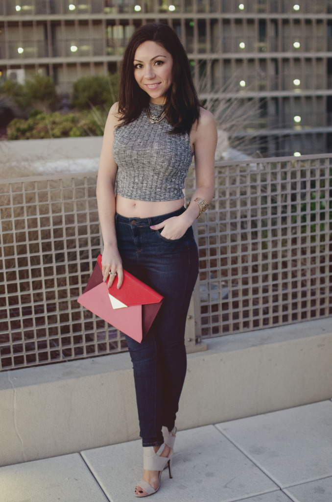 Blogger Nihan smiling and posing to show her edgy cool and sexy outfit; a gray top, blue jeans, leather jacket, neutral high-heels and a red and pink clutch.